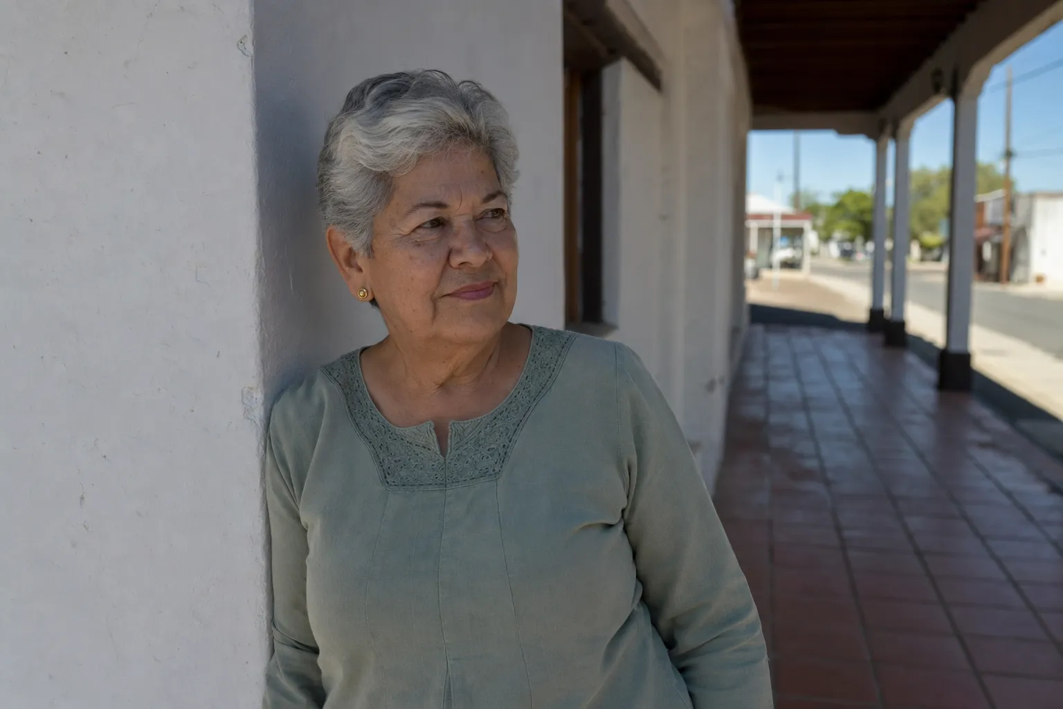 An older woman with white hair standing in the shade beside a whitewashed column, looking out with a soft smile