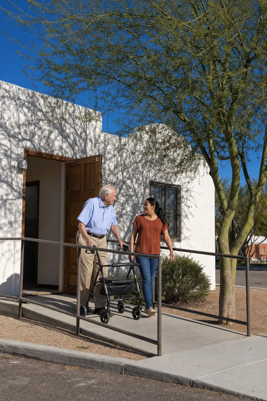 An older man with a walker and a young woman talking on the ramp outside the open front door of a whitewashed building, a palo verde beside them